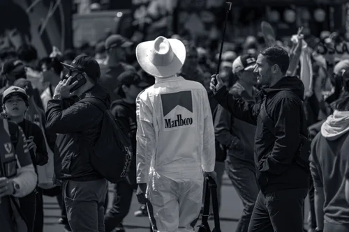 Black and white photo of a fan in vintage white Marlboro racing overalls and a wide-brimmed hat, seen from behind in the crowd