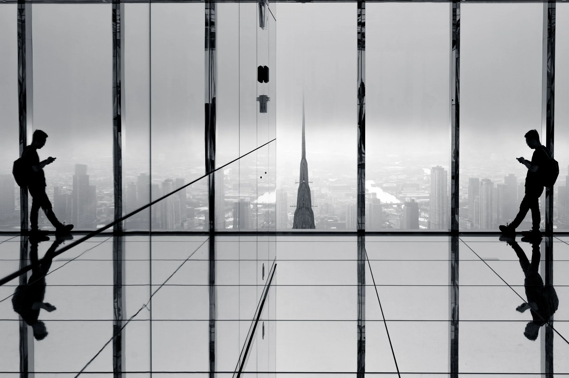 A man on his phone silhouetted against floor to ceiling glass and reflected symmetrically on both sides of the mirrored room at Summit One Vanderbilt, the Chrysler Building spire in fog beyond, New York, in black and white.