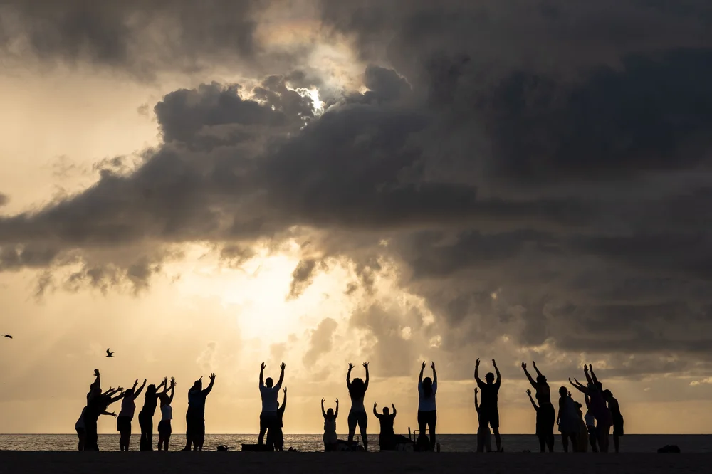 A line of people silhouetted at the water's edge with arms raised, dramatic clouds and golden light behind, Miami Beach.