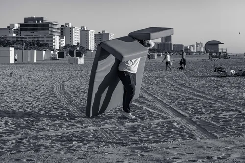 A person on the sand wrestling a large windblown canopy, the Miami Beach skyline behind, in black and white.
