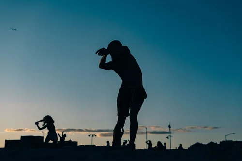 A dancer silhouetted against a deep blue dusk sky, a second figure crouched nearby along the low horizon at Rockaway Beach, New York.