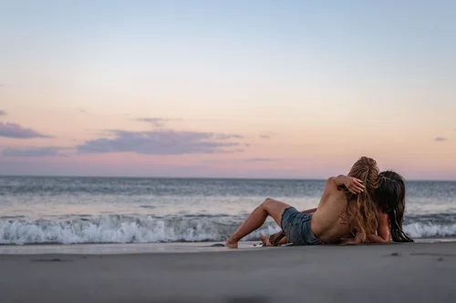 A couple reclining together on the sand at the water's edge beneath a soft pink dusk sky at Rockaway Beach, New York.