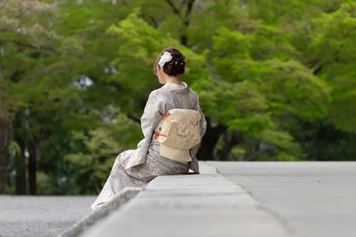A person in kimono seated on a low wall facing a green garden, seen from behind, Kyoto.