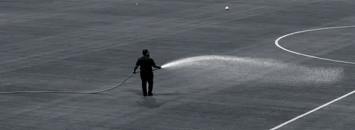 Black and white photo of a lone groundskeeper watering a sports pitch with a hose, the field's white line markings curving across the frame