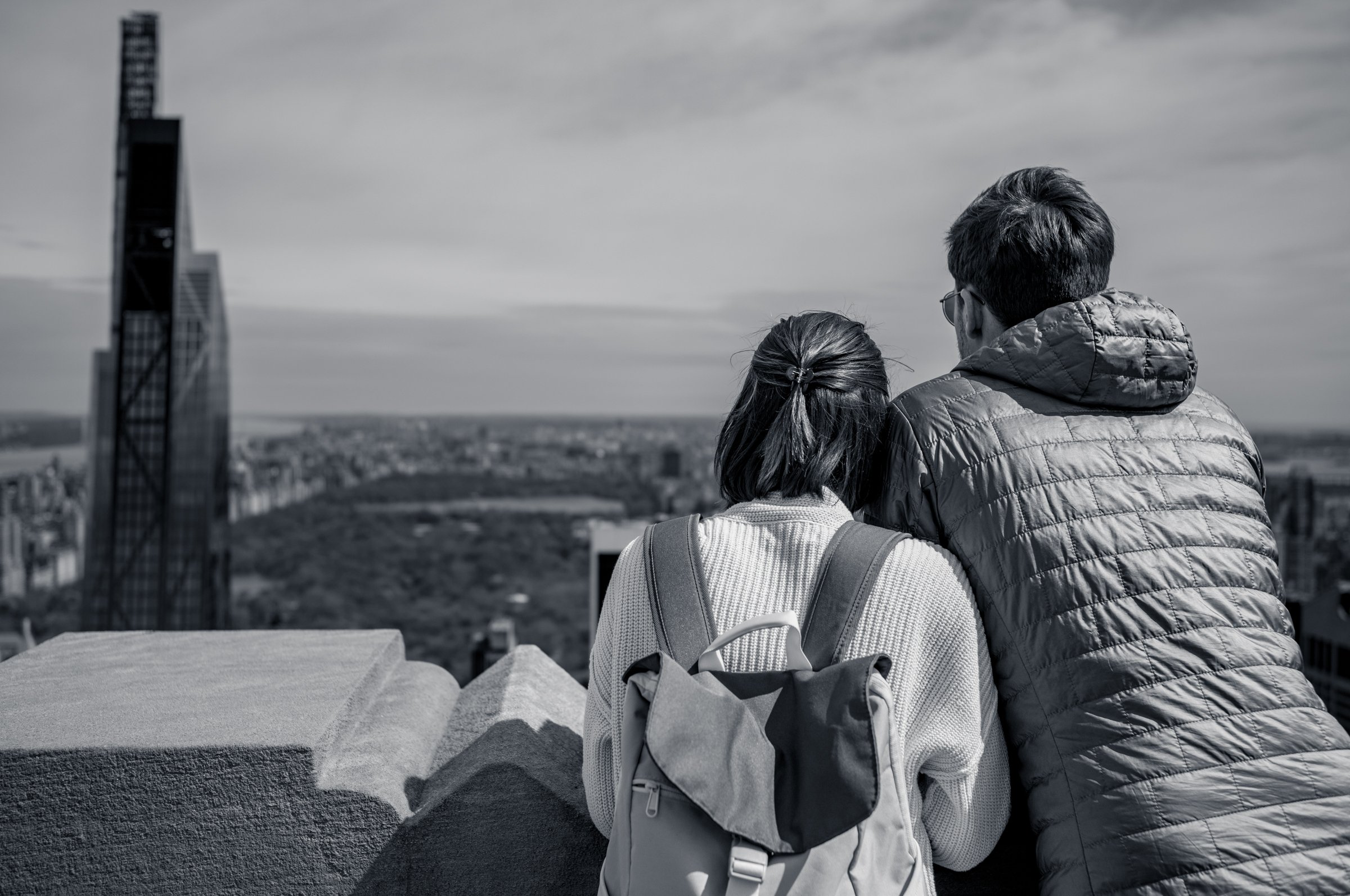 A couple seen from behind leaning on the concrete parapet on the north side of the Top of the Rock observation deck, Central Park spread out grey-green below them, in black and white.