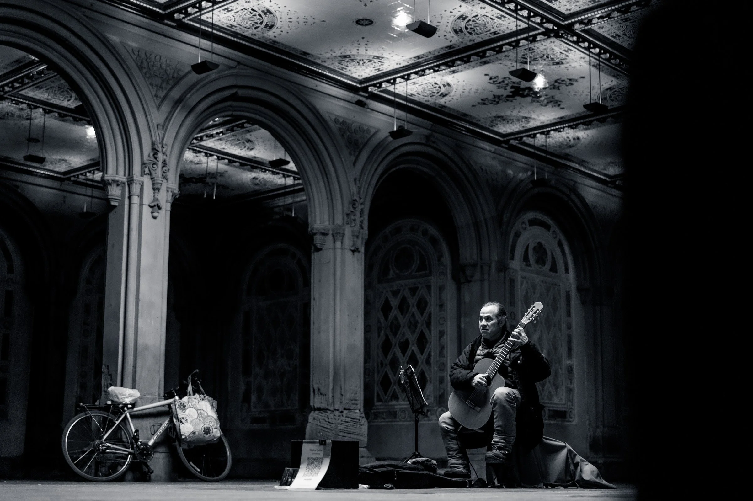 Classical guitarist Carlos König seated playing beneath the tiled arcade ceiling of Bethesda Terrace in Central Park, a bicycle propped against a column, New York, in black and white.