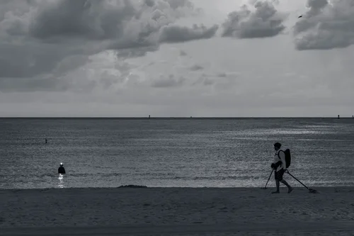 Two figures walking along the shoreline under a cloudy sky on a wide open beach, Miami Beach, in black and white.