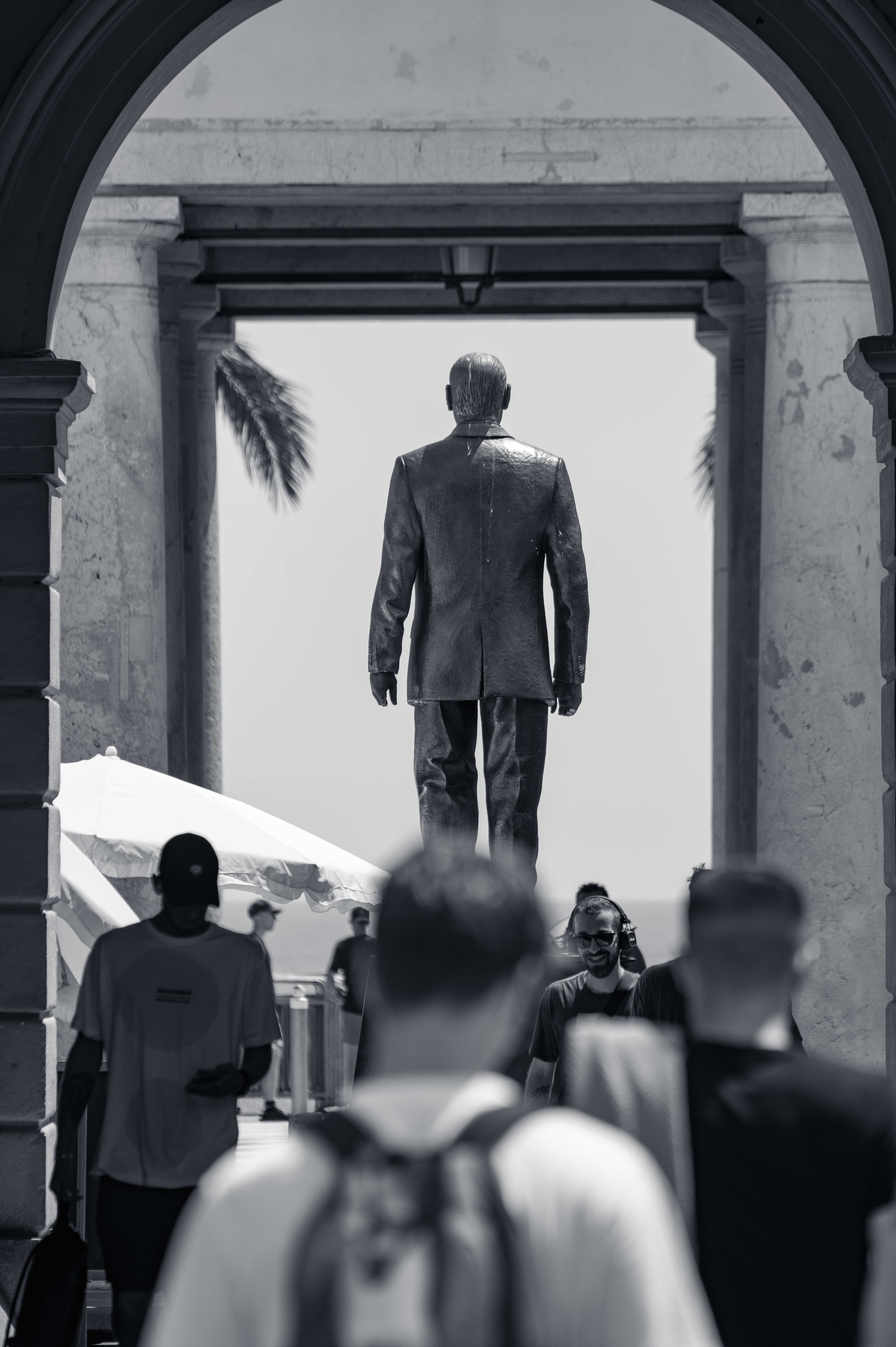 A bronze statue of a suited man on a plinth, seen from behind through a stone archway, with a crowd passing in front in Vieux Nice.