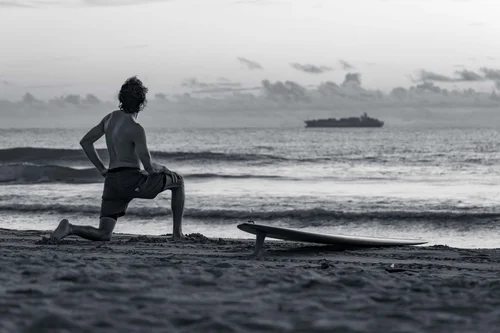 A surfer kneeling beside a board at the water's edge, ships on the horizon, Miami Beach, in black and white.