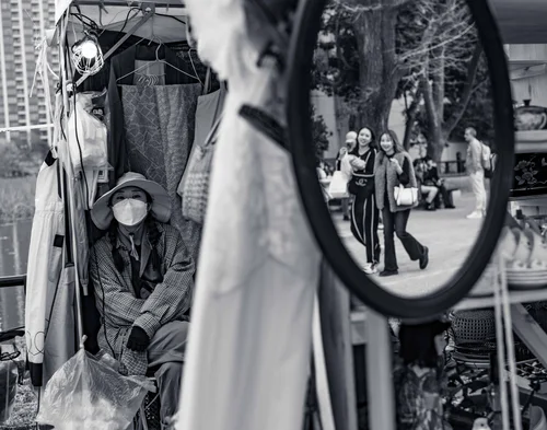 A street scene reflected in a round convex mirror, a masked vendor seated alongside, Ueno Park, Tokyo, in black and white.