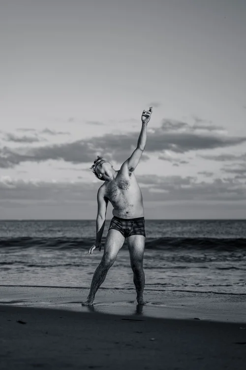 A dancer standing on the sand with one arm raised high toward the sky, the sea behind at Rockaway Beach, New York, in black and white.