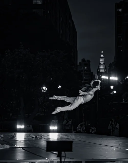 A dancer suspended in a horizontal mid-air leap across a dark plaza, city lights and buildings behind at night in Battery Park, New York, in black and white.