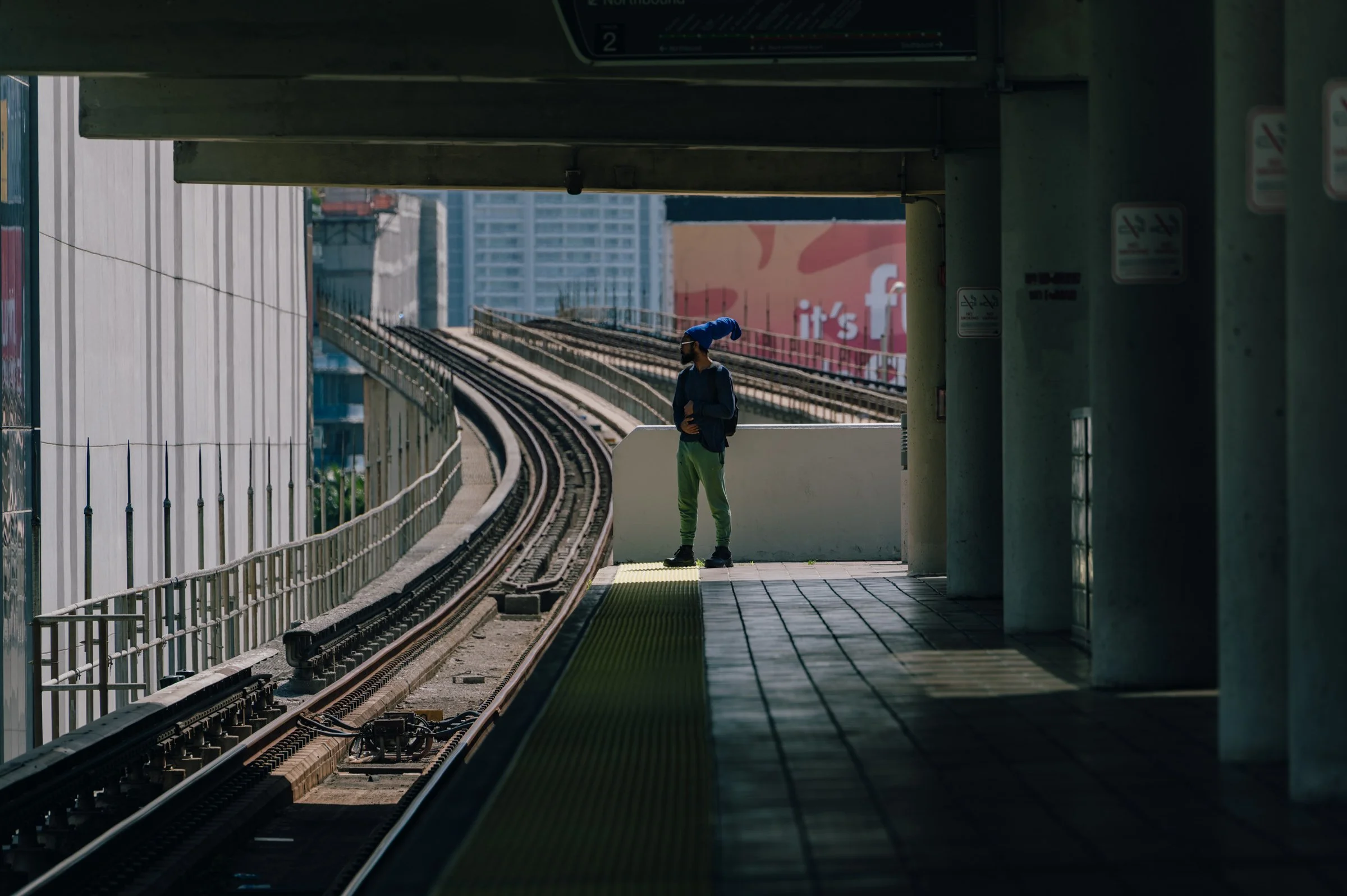 A single passenger waiting on an elevated transit platform in Miami, the track curving away to the left and the city towers beyond.