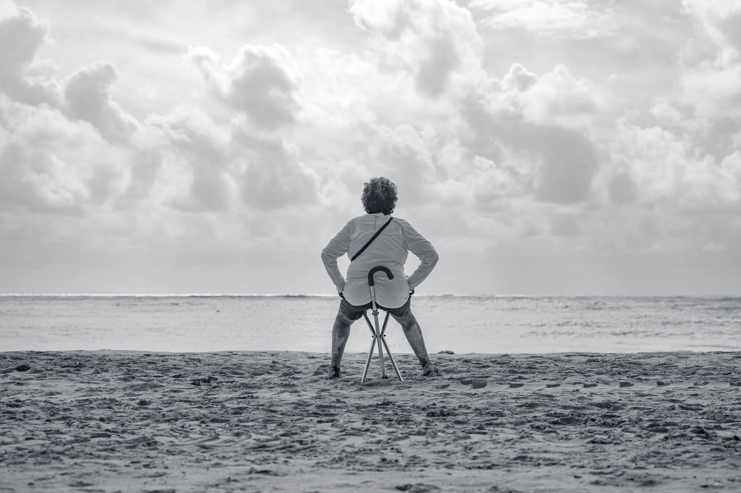 A woman sitting on a small folding stool facing the sea at Playa del Carmen, hands on her hips, a walking cane hooked over the frame beside her, in black and white.