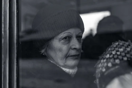 A close black and white portrait of an older woman in a knitted hat, seen past a window frame, her gaze turned aside, Bucovina, Romania.