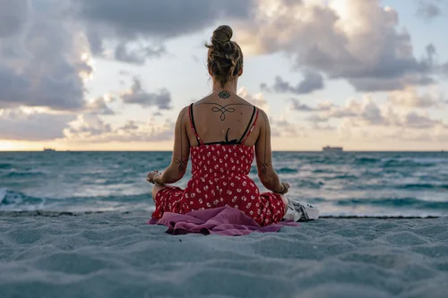 A woman in a red dress sitting cross-legged on the sand facing the sea, seen from behind beneath a soft sky, Miami Beach.