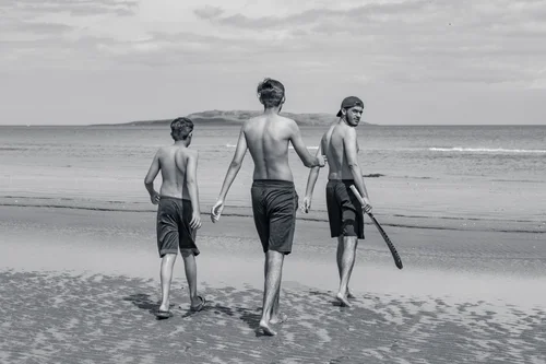 Three young men walking away toward the sea across the wide sand at Portmarnock beach, Dublin, in black and white, one carrying a stick at his side.