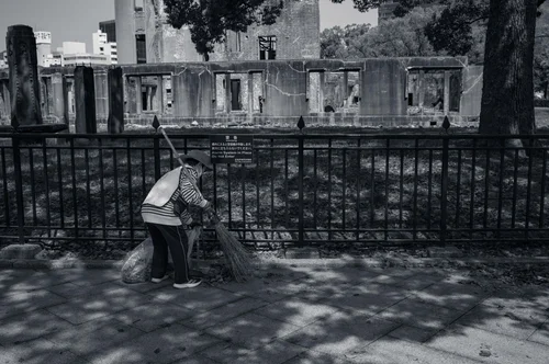 A person sweeping in dappled light before a fenced building, Hiroshima, in black and white.