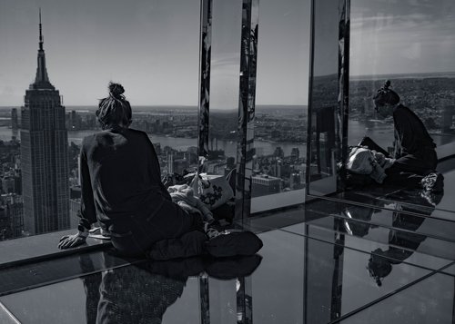 Two people seated at the floor-to-ceiling windows of a New York viewing gallery, the Empire State Building rising through the glass and mirrored in the polished floor around them.