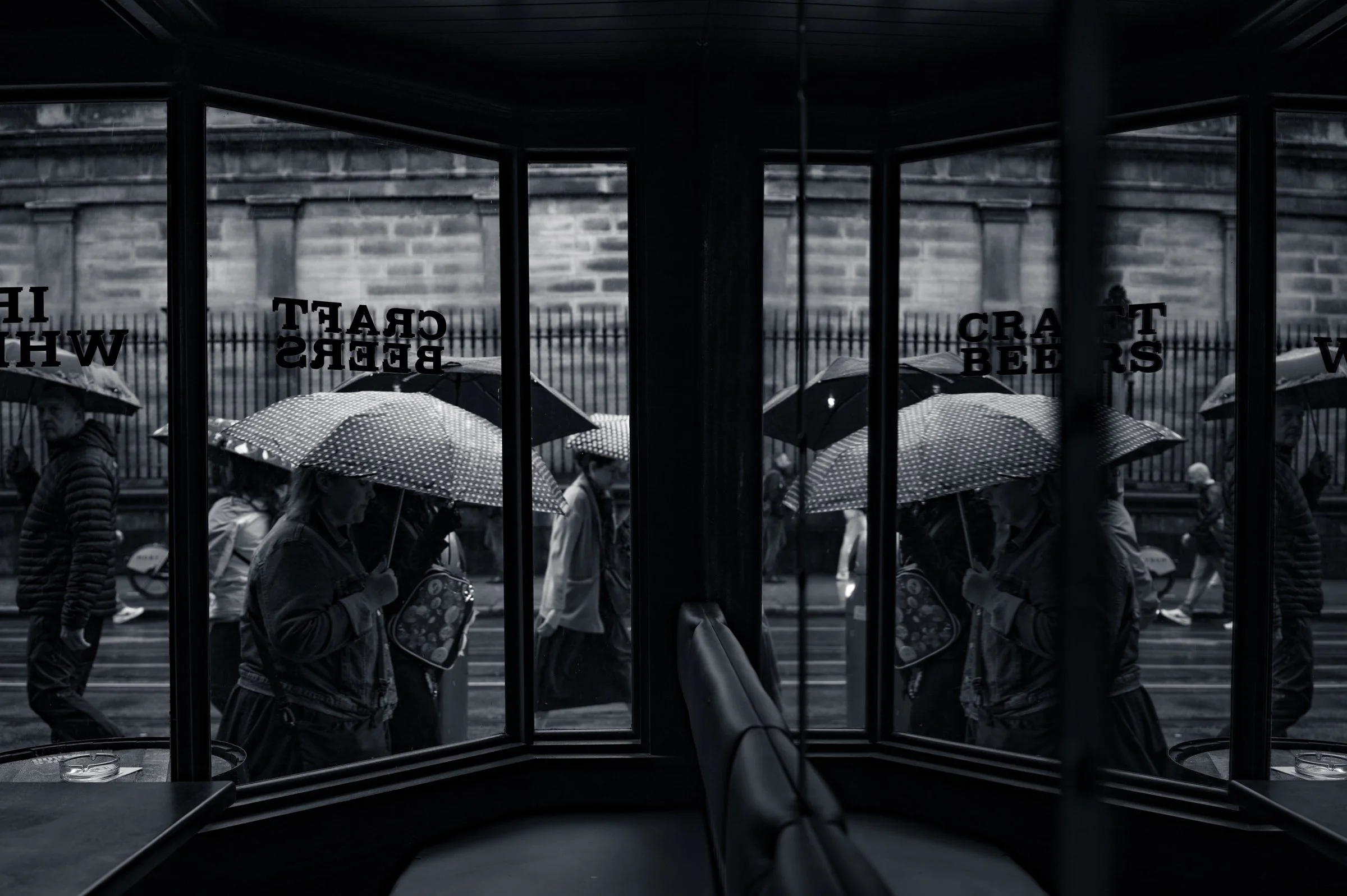 Figures under umbrellas seen through the mirrored glass panels of a shelter, their reflections doubled against the street beyond.