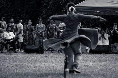 A dancer spinning on the grass in a dark robe, the fabric flaring out, a crowd watching from the treeline at Astoria Park, New York, in black and white.