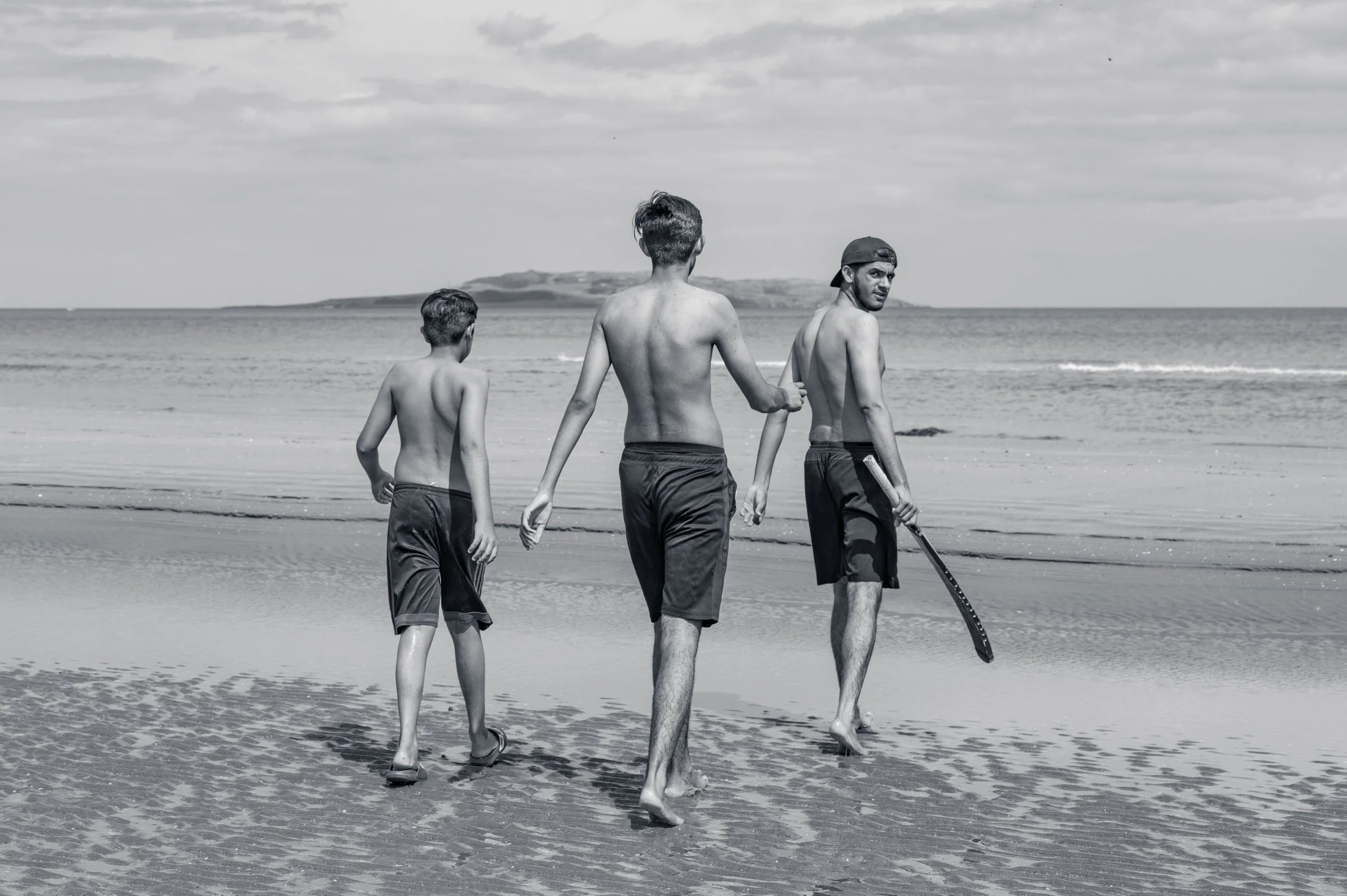 Three young lads in swim shorts walking away from the camera down a wide empty strand towards the sea, in black and white, a headland in the distance.