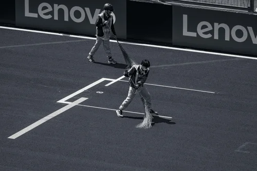 Black and white photo of two track marshals sweeping a race circuit with long brooms beside advertising hoardings