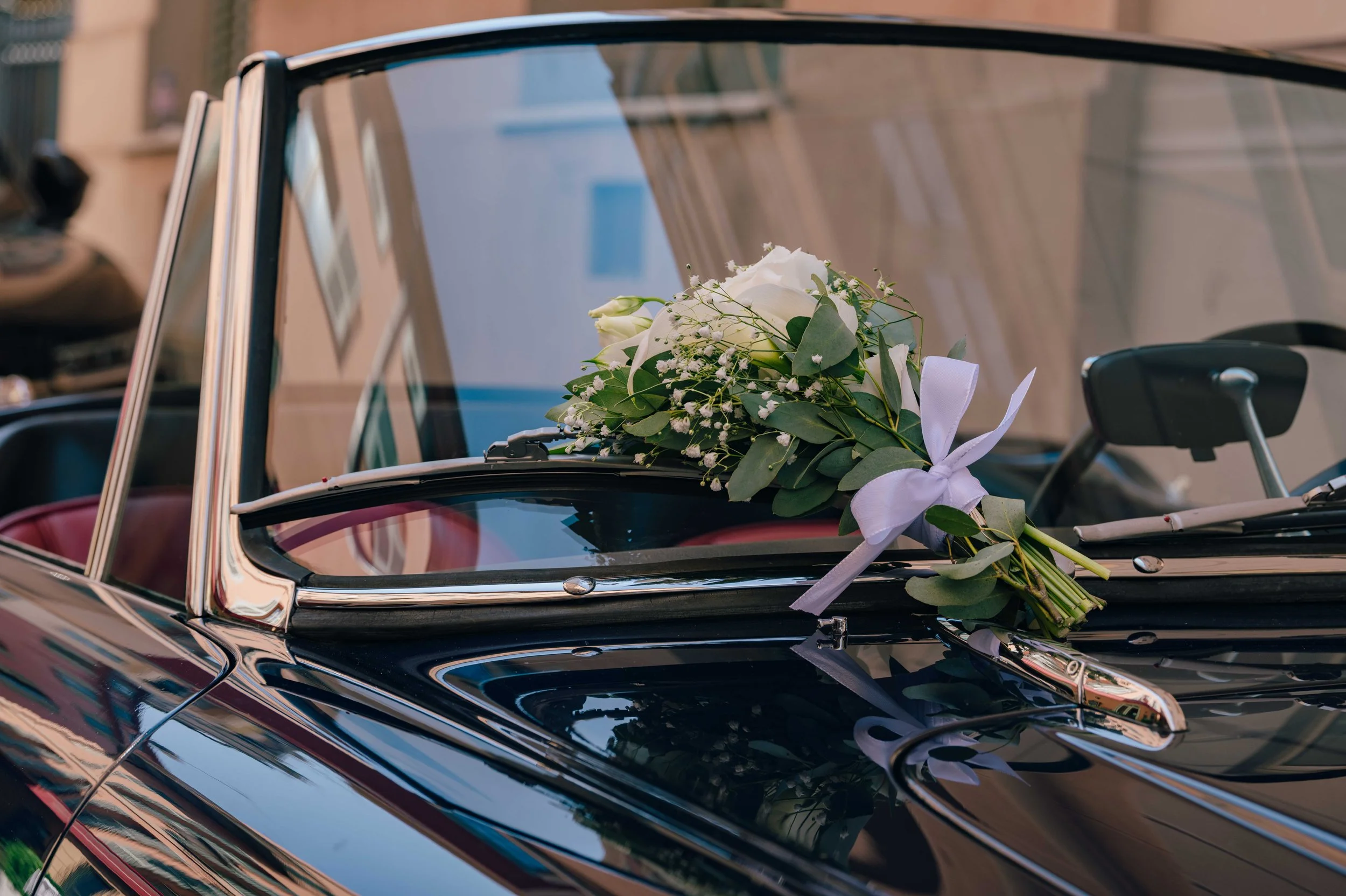 A white bridal bouquet tied with ribbon resting on the boot of a dark vintage convertible wedding car in the streets of Nice.