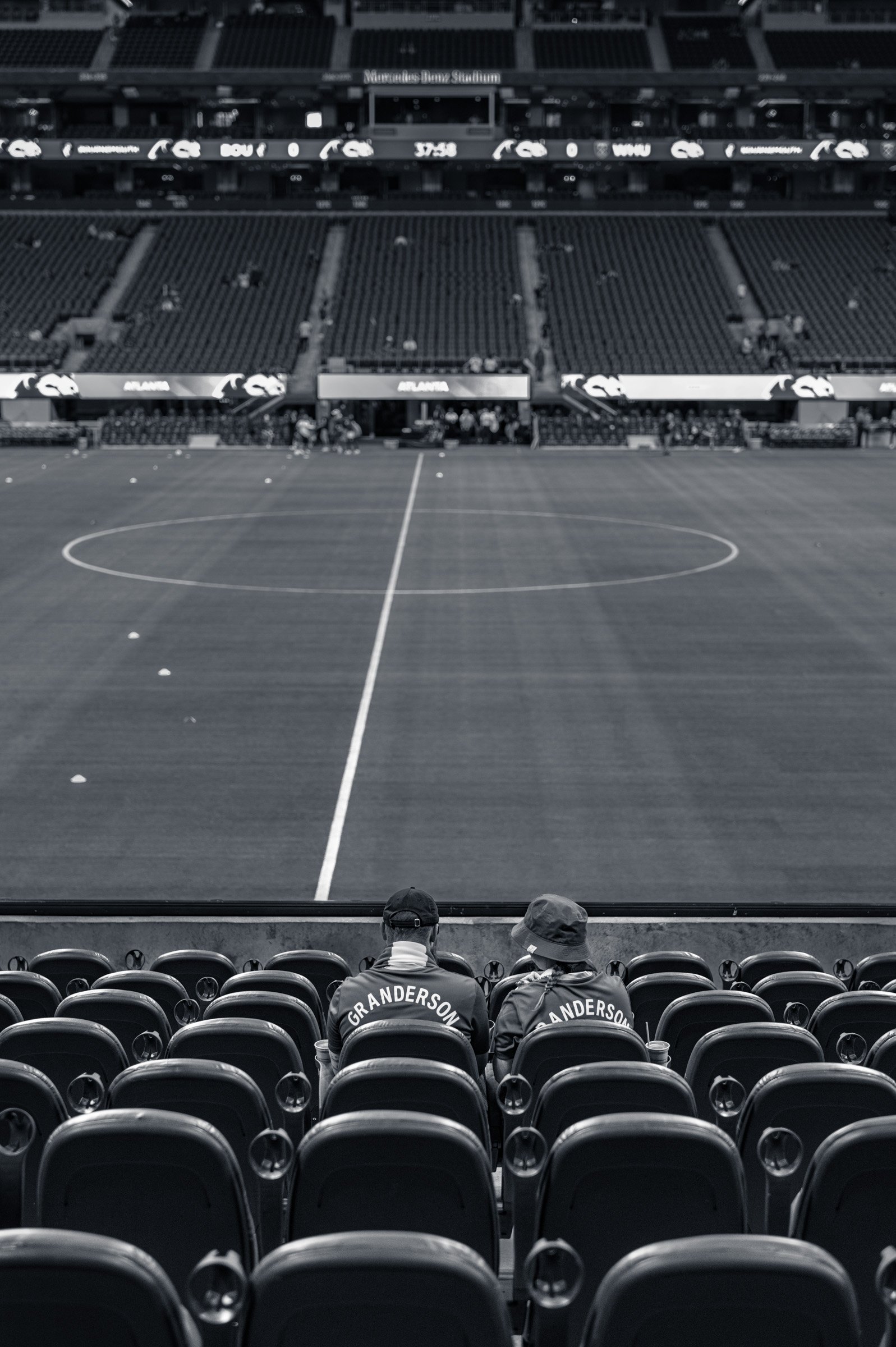 Two supporters sitting alone in an empty tier at Mercedes-Benz Stadium in Atlanta well before kick-off, the pitch and the far stands completely empty, in black and white.