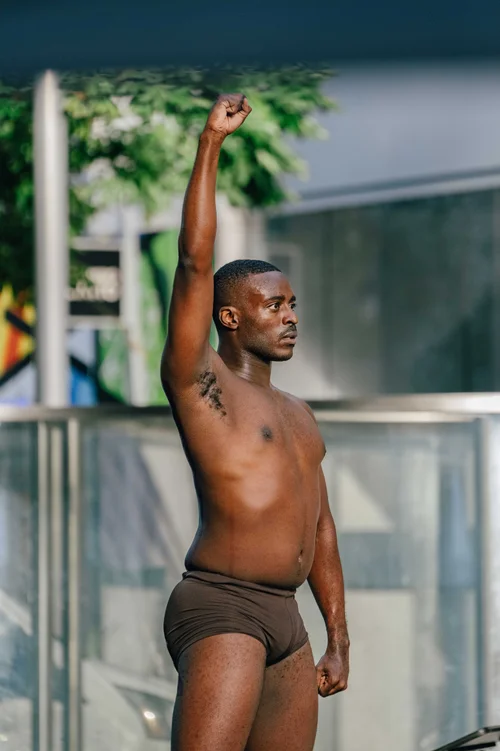 A dancer standing with one arm raised straight overhead in front of a glass structure edged with greenery, Miami.