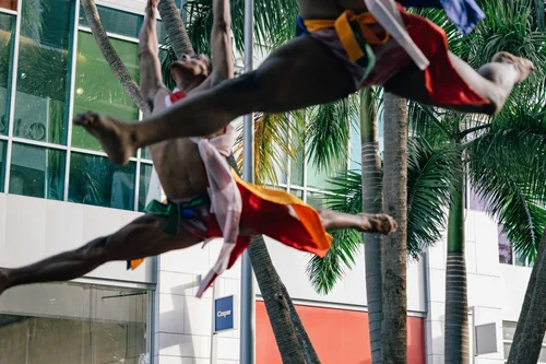 Two dancers suspended in a mid-air leap in bright garments, palm trees and a glass building behind, Miami.