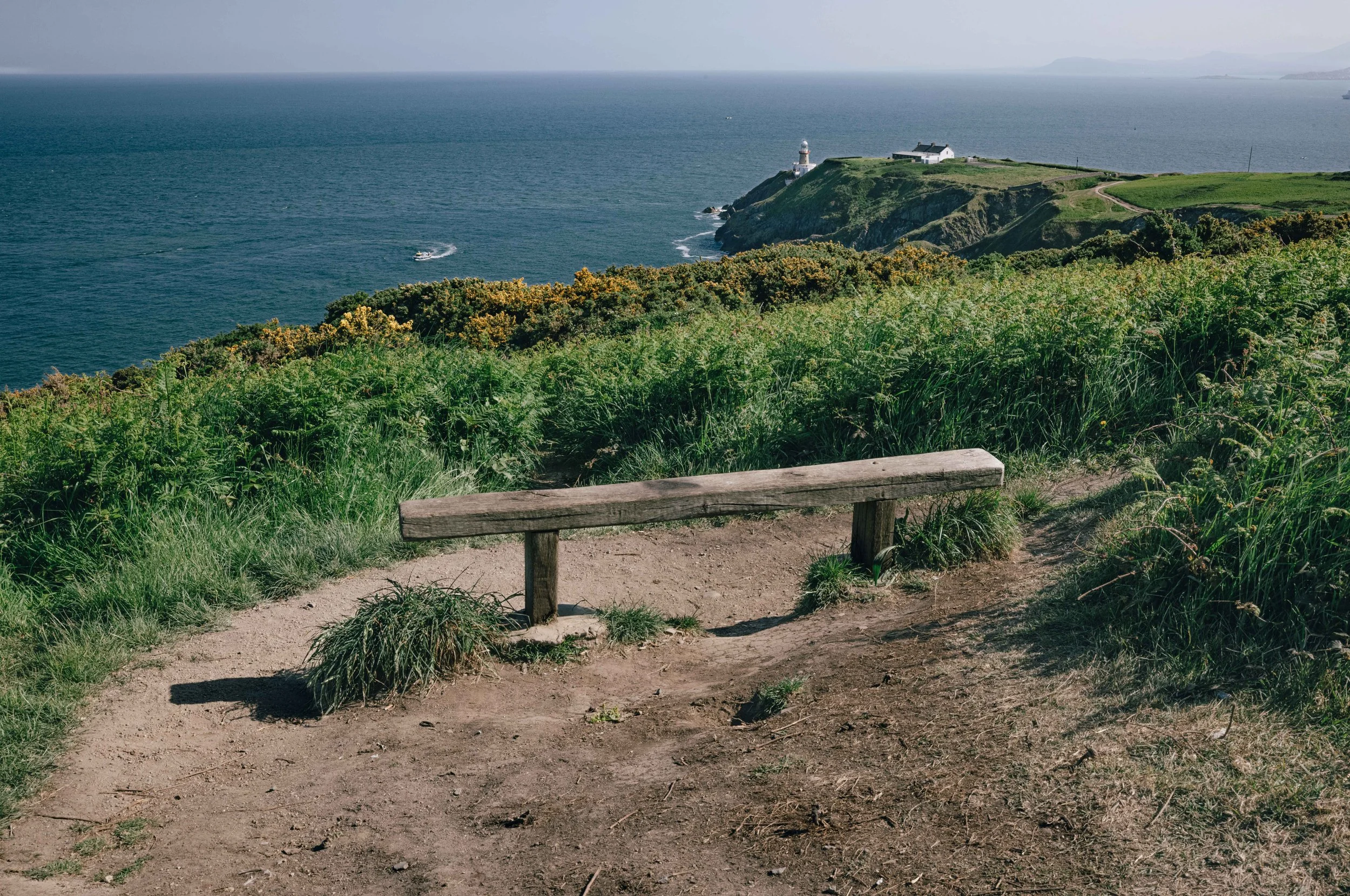 A weathered wooden bench overlooks the cliffs at Howth Head, with the Baily Lighthouse and the Irish Sea stretching into the distance.
