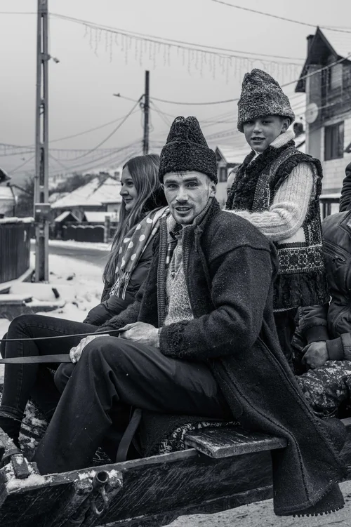 Two men in tall fur hats seated side by side at a winter market, a village behind under a web of wires, Bucovina, Romania, in black and white.