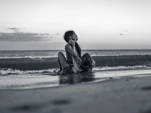 A figure sitting on the wet sand at the shoreline with knees drawn up, facing the sea at Rockaway Beach, New York, in black and white.