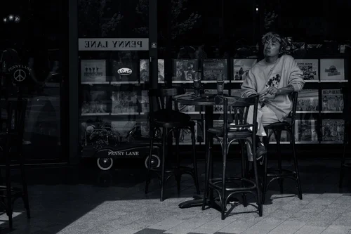 A person sitting alone at a small table outside a lit shopfront at night, Tokyo, in black and white.