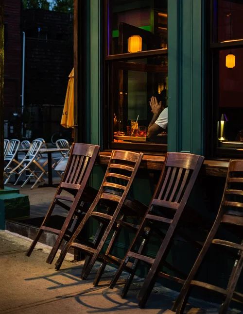 Wooden chairs stacked outside a warmly lit bar at night, a lone patron seated inside, in New York.