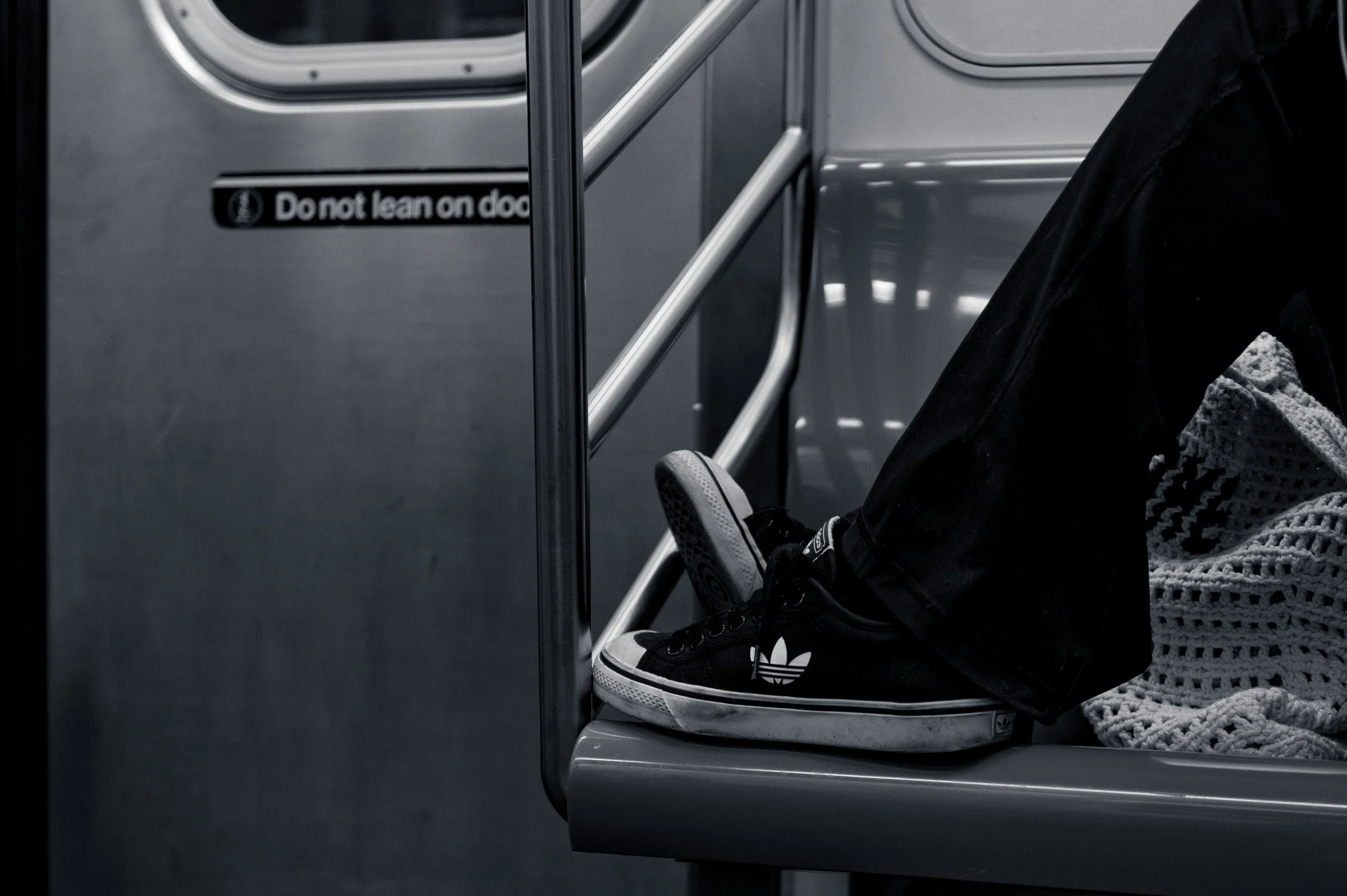 Subway car interior with a person's legs stretched out and Adidas trainers resting on the seat edge beside a crocheted bag, a Do not lean on door sign behind, in black and white.