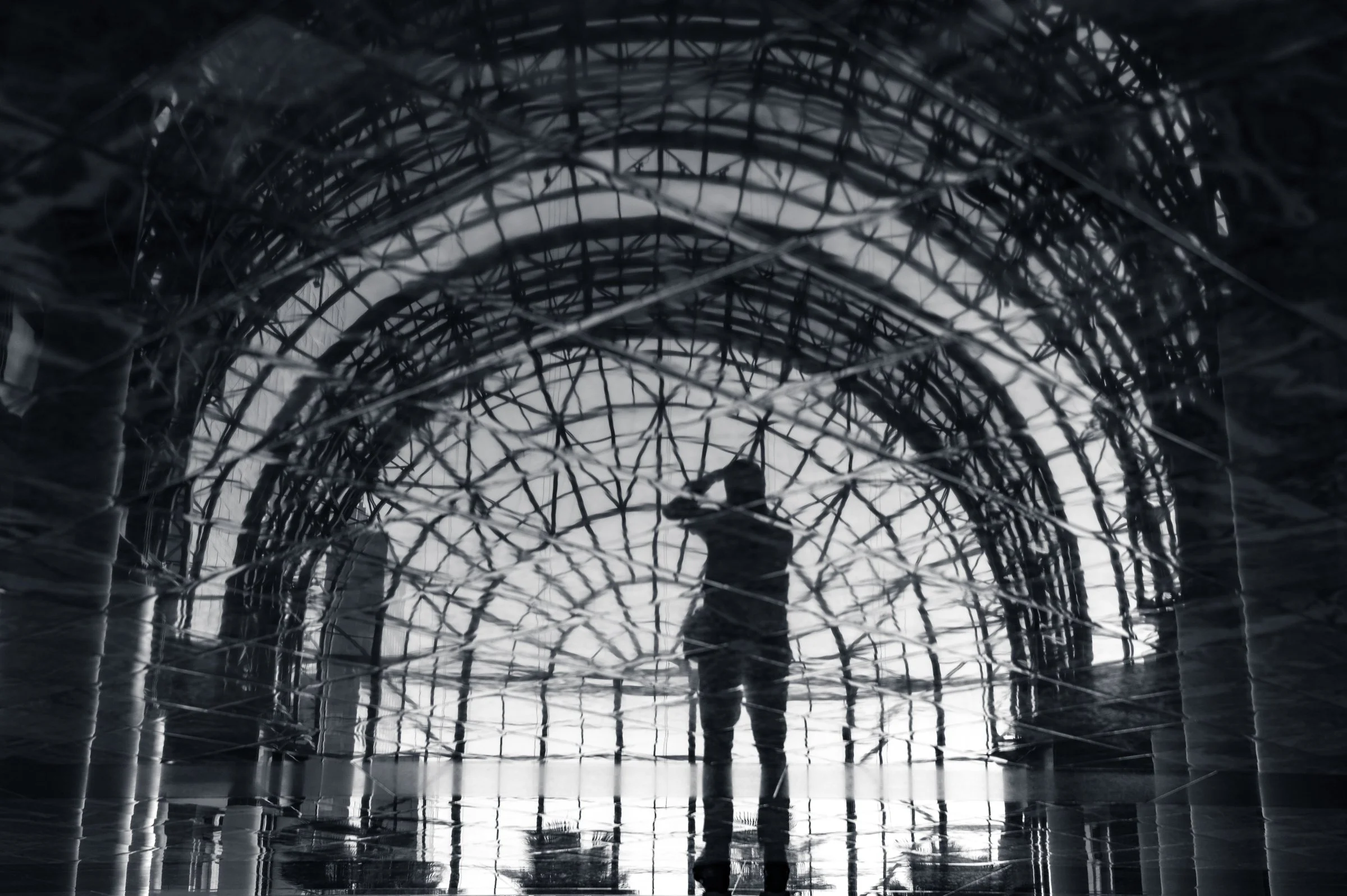 A lone figure with a camera raised to their face standing at the centre of a huge vaulted hall of arched steel ribs, the whole picture broken into ripples and doubled by a wet reflective floor, in black and white.