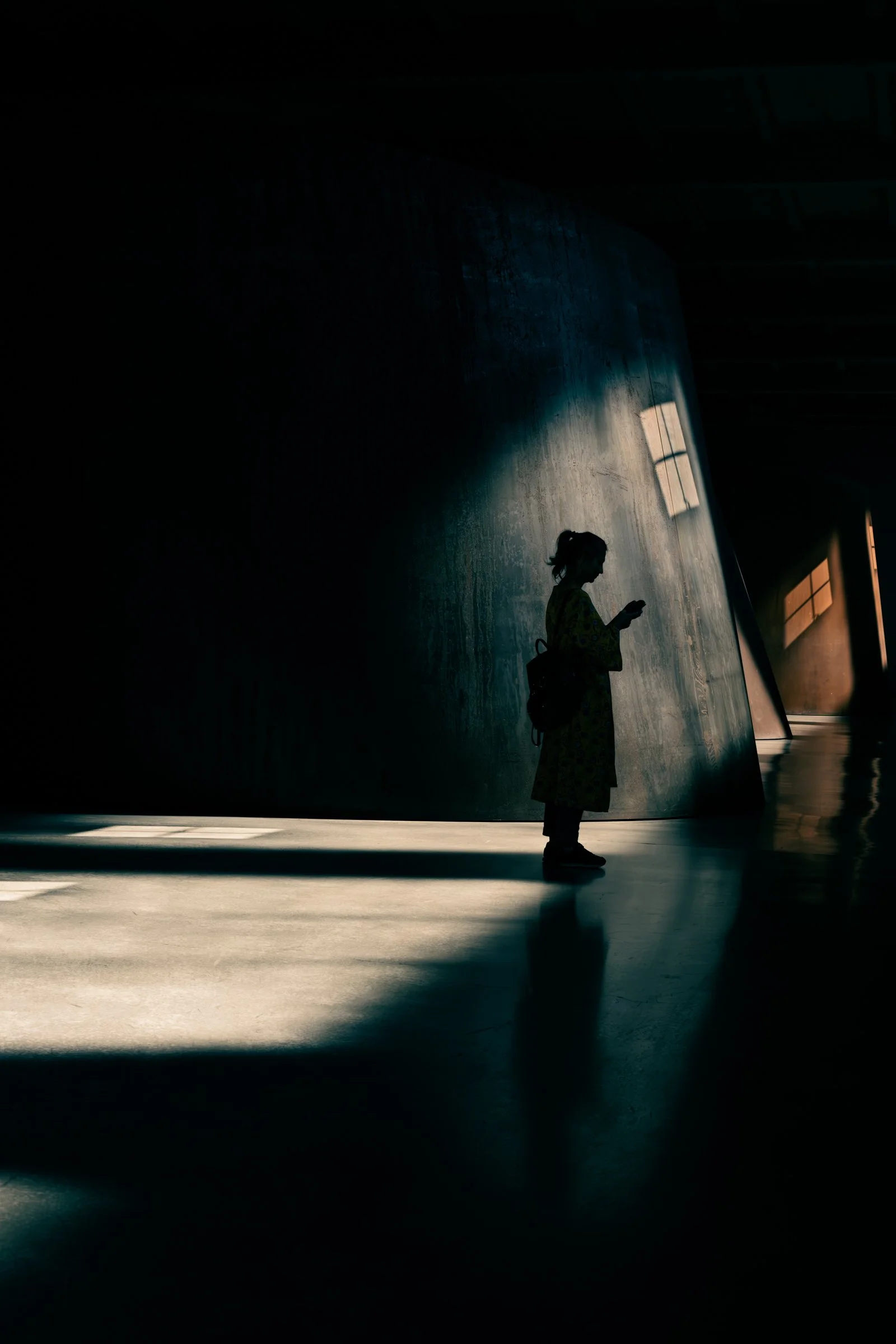 A woman standing in a shaft of daylight against a large concrete wall at Dia Beacon, phone in hand, the rest of the room in shadow.