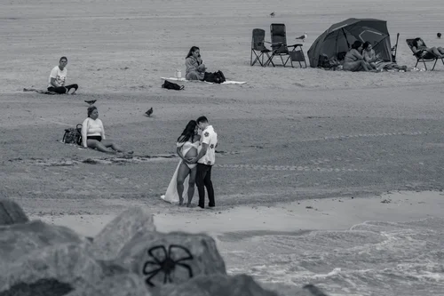 Beachgoers scattered across the sand with chairs and umbrellas, a couple standing close in the middle, Miami Beach, in black and white.