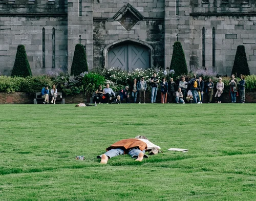 A person lying alone on the grass in the grounds of Dublin Castle, a crowd gathered by the Gothic stone facade behind.
