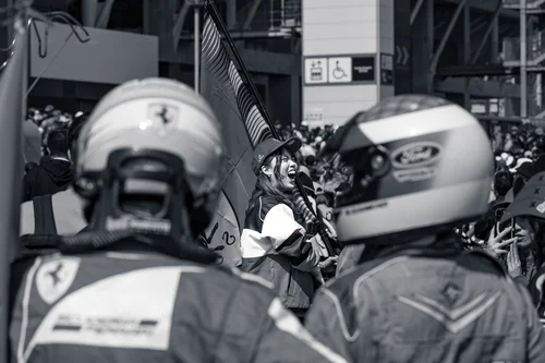 Black and white photo of two fans in Formula 1 racing helmets and team overalls seen from behind, with another fan cheering and waving a flag between them