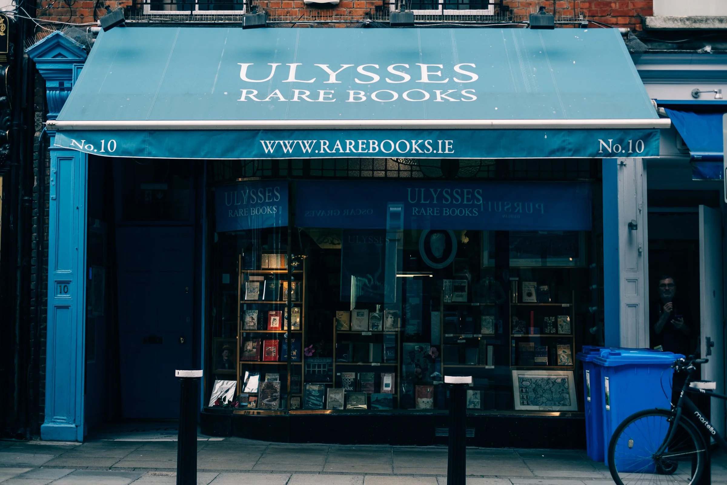 The blue-painted shopfront of Ulysses Rare Books in Dublin, its window packed with secondhand books, a bicycle and a blue recycling bin on the pavement outside.