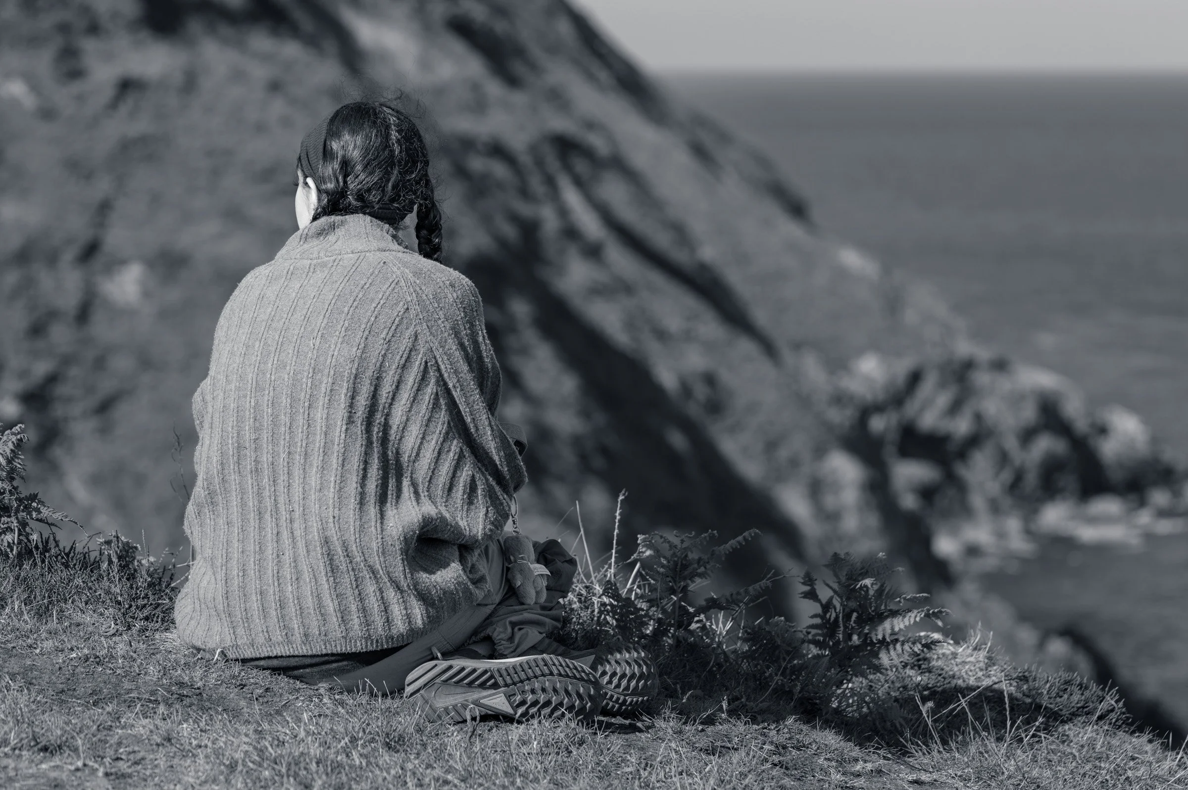 A woman seen from behind sitting at the cliff edge on Howth Head with her hair plaited, the cliff face and open water beyond her, in black and white.