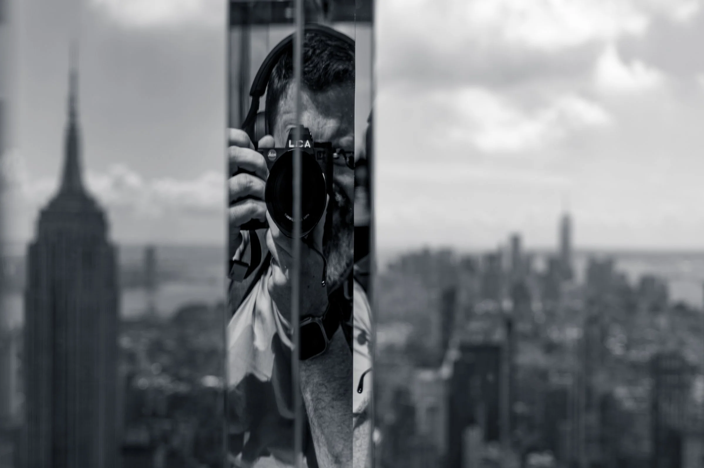 The photographer Paul O'Malley reflected in a narrow mirrored column with a Leica raised to his eye, the Empire State Building and the Midtown skyline visible either side, at Summit One Vanderbilt, New York, in black and white.