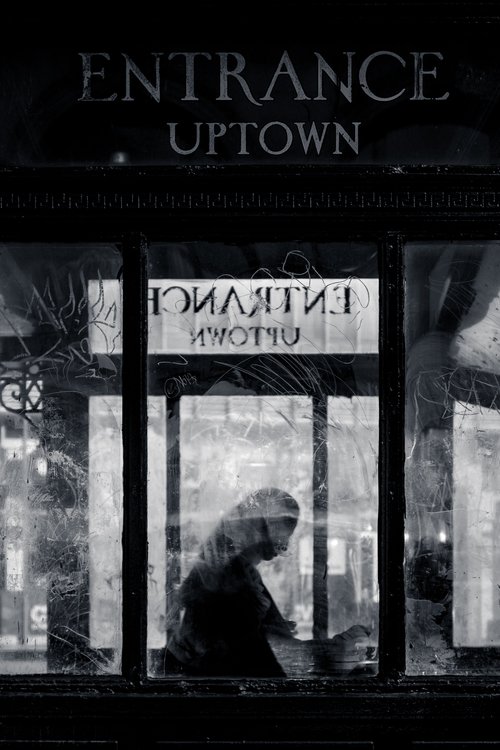 A lone figure behind glass, with reflected 'Entrance' and 'Uptown' signage marking a New York subway entrance in black and white.