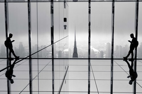 Two silhouetted figures at the floor-to-ceiling windows of a high-rise viewing gallery in New York, their reflections mirrored in the polished floor as the city stretches out below.