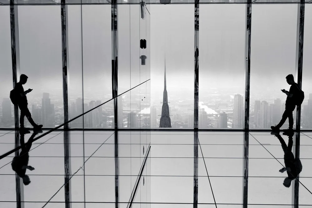Two silhouetted figures at the floor-to-ceiling windows of a high-rise viewing gallery in New York, their reflections mirrored in the polished floor as the city stretches out below.