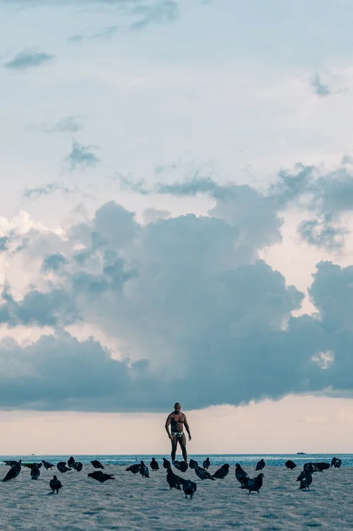 A solitary figure standing in the shallows beneath a wide cloudy sky, birds scattered on the water, Miami Beach.