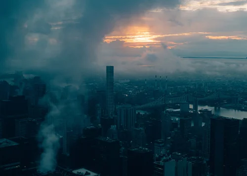 Dark aerial view of a city skyline at dawn, orange light breaking through heavy clouds with smoke rising over the buildings and a bridge crossing the river below.